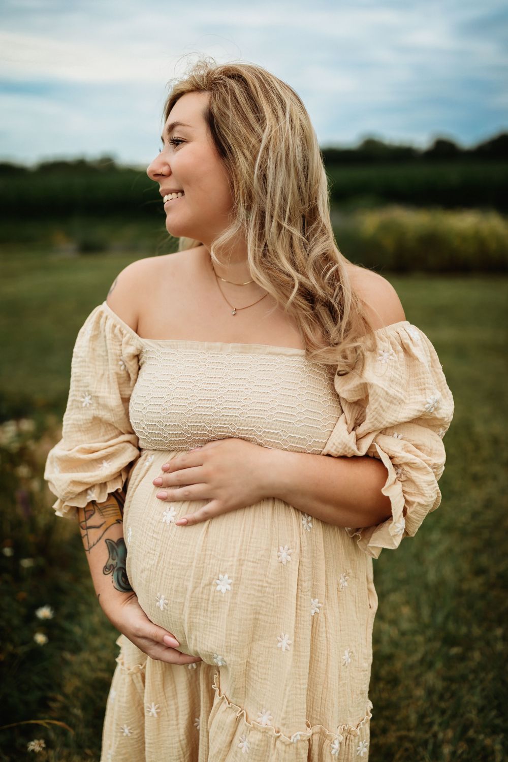 A person in a cream-colored vintage dress stands in a sunny field, showcasing a romantic, ethereal maternity photoshoot.