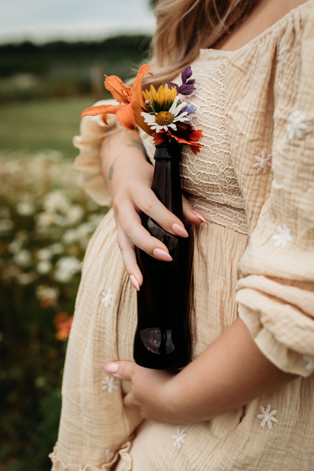 Close-up shots of hands holding a dark wine bottle with orange flowers against a cream dress in a garden setting.