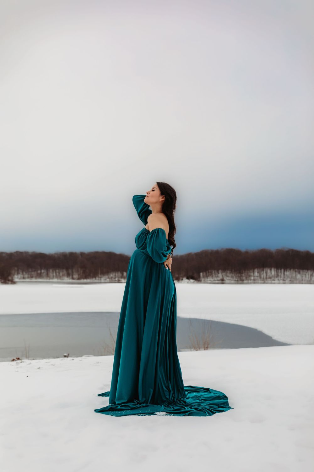 A person in a flowing teal gown poses gracefully against a snowy winter landscape with trees in the background.