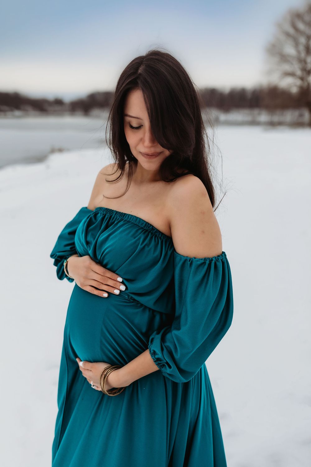 A pregnant woman in a teal off-shoulder dress poses outdoors in a snowy winter landscape.