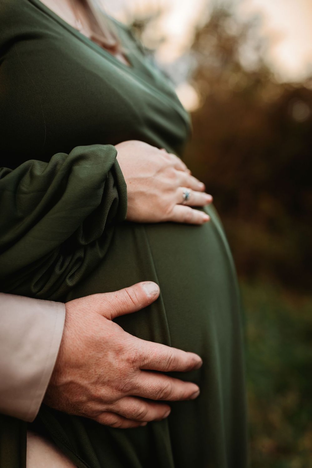 Hands lovingly cradling a pregnant belly in an olive green dress at sunset.