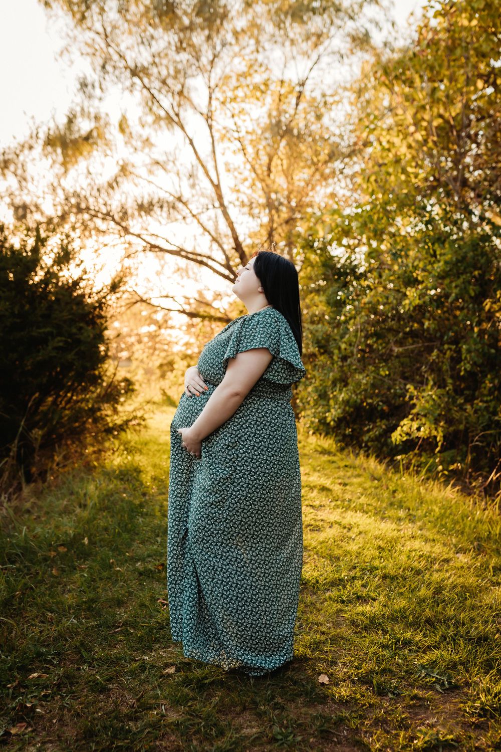 A pregnant woman in a green polka dot dress sits on a grassy hill during golden hour sunset.