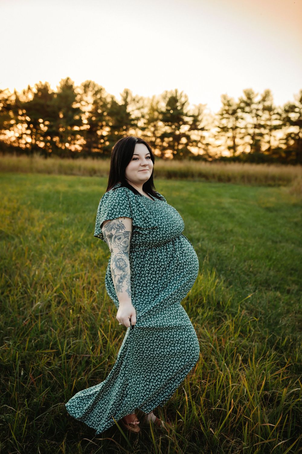 A person in a green floral dress twirls in a grassy field at sunset while doing a photoshoot.