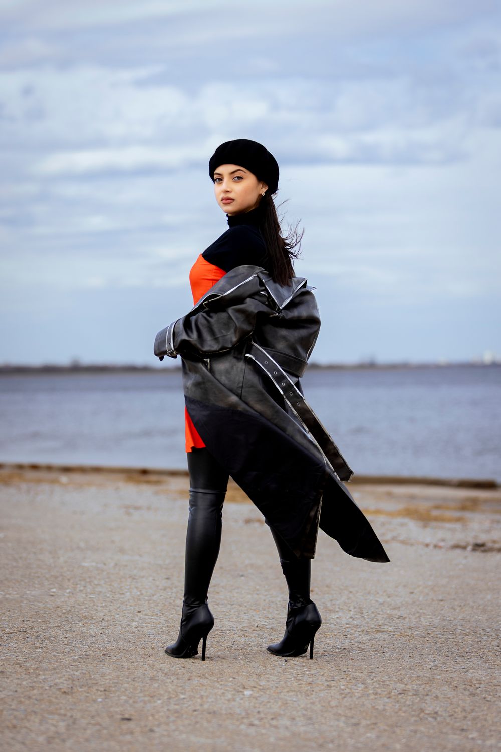 Fashionable figure in black beret and boots poses dramatically with flowing coat on cloudy beach shoreline.