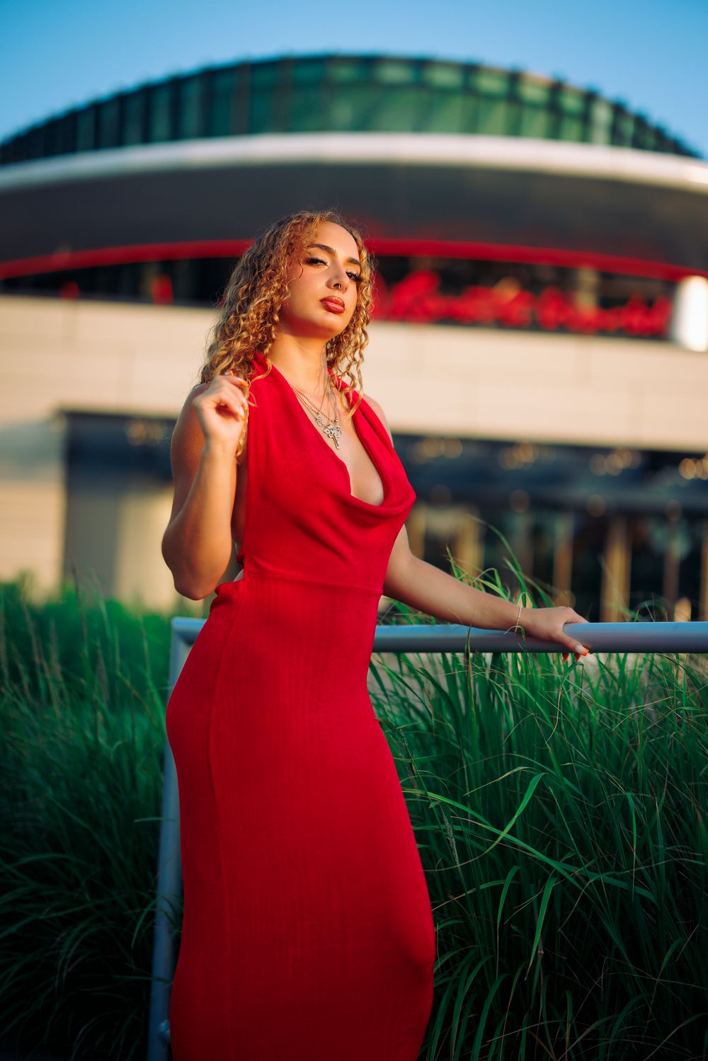 A person in a red dress poses gracefully in tall grass at sunset with modern architecture in the background.
