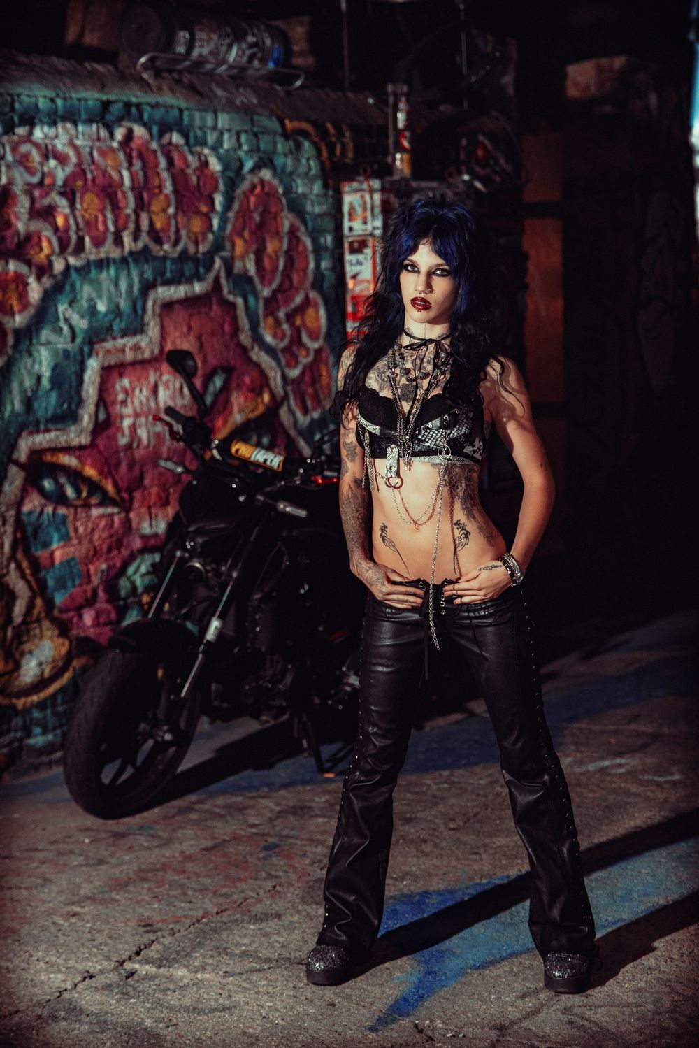 Edgy fashion photo series against graffiti wall shows model in black leather pants with motorcycle in dark urban setting.