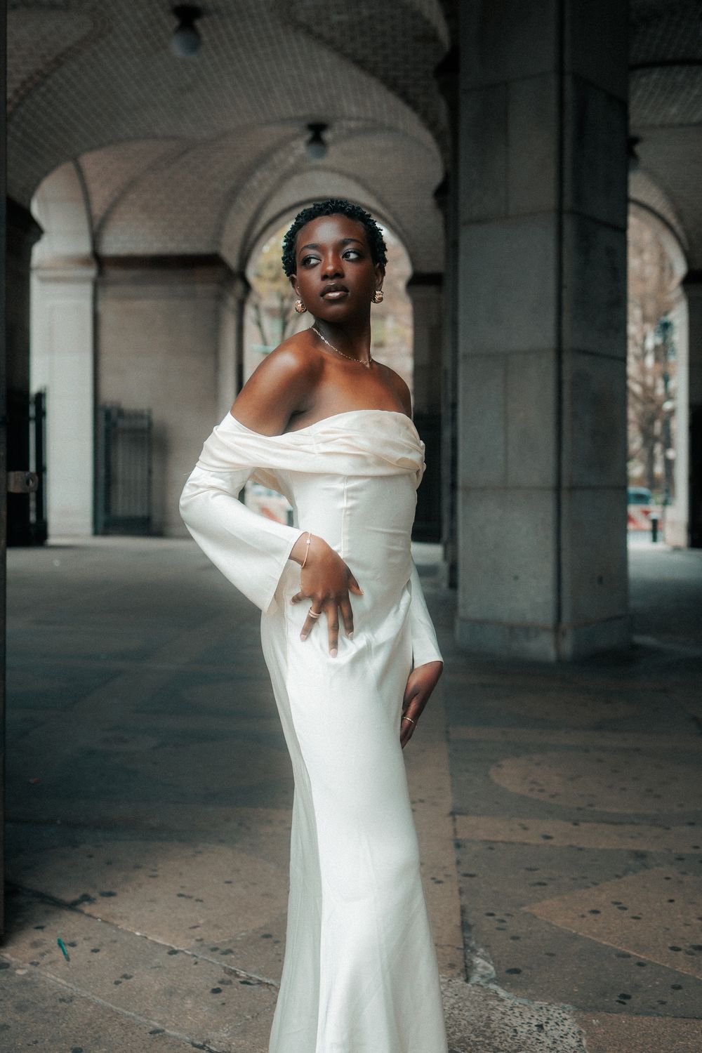 A person in a flowing white off-shoulder dress poses dramatically in an industrial concrete space.