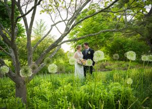 A couple stands in a lush green garden adorned with white dandelion-like spherical decorations hanging from trees.