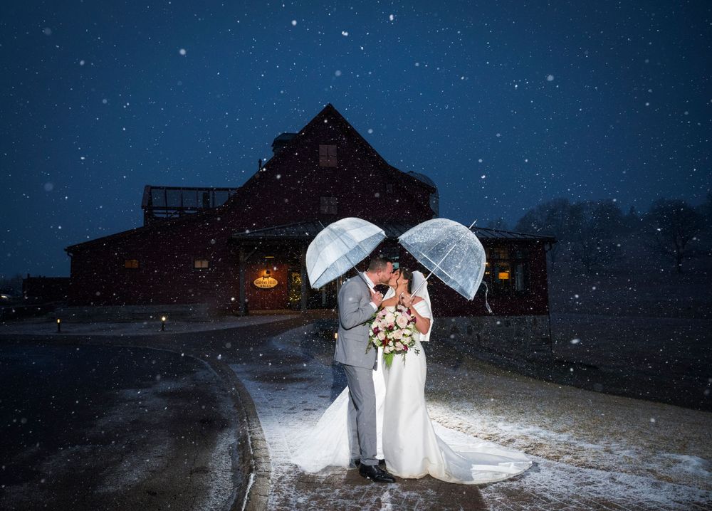 Romantic winter wedding photo with snowfall outside a rustic barn venue.
