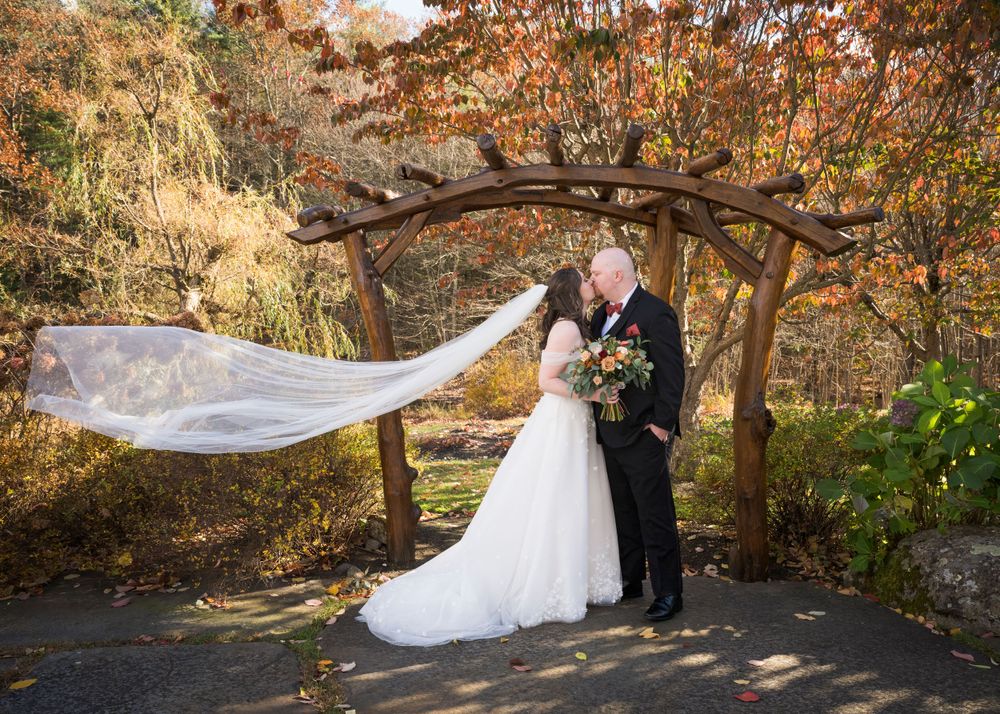 A flowing wedding veil catches the autumn breeze under a rustic wooden arch in a garden setting.