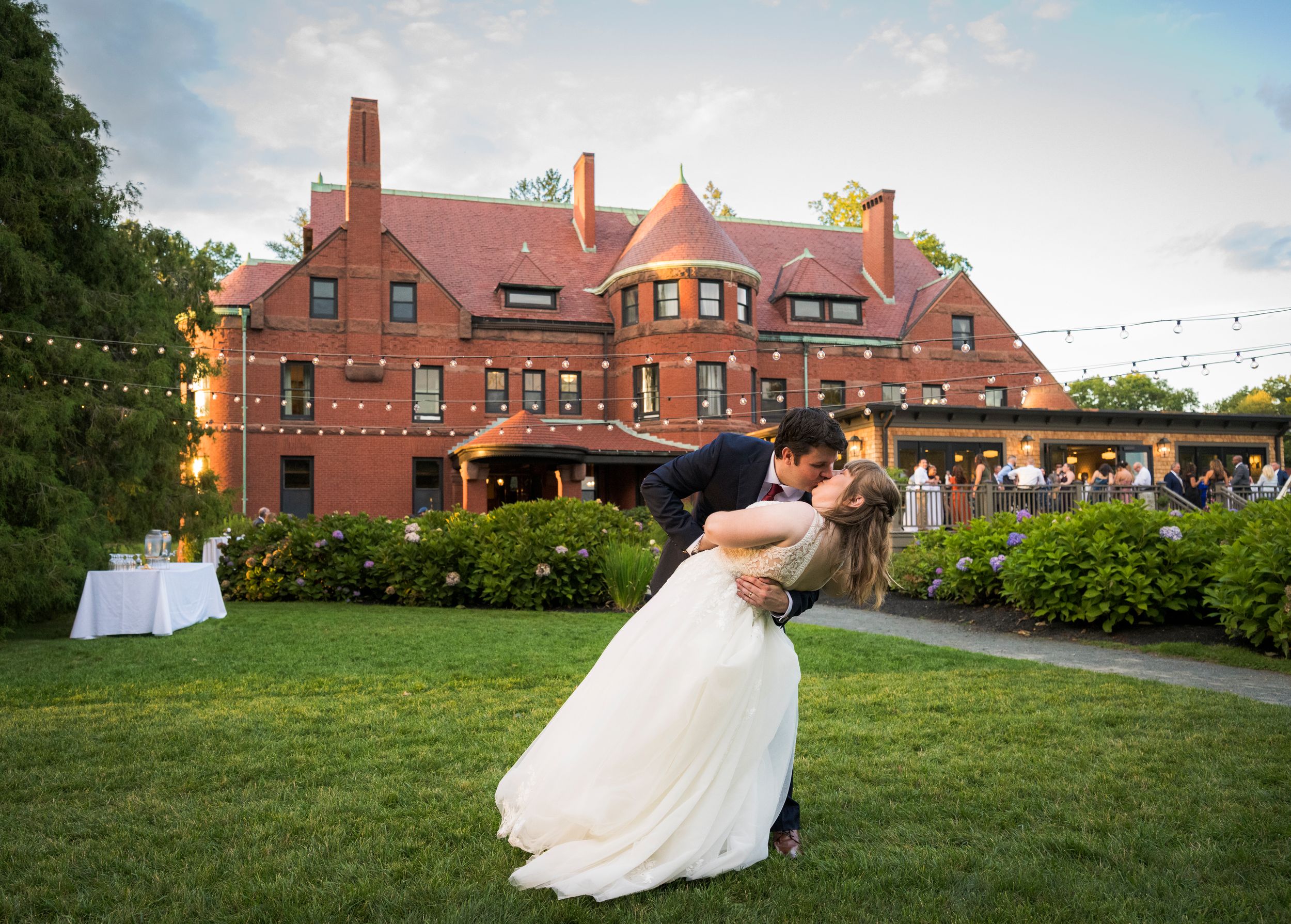 Bride in a white gown and groom share a dramatic dip kiss on a lush green lawn in front of a historic red brick mansion at sunset.
