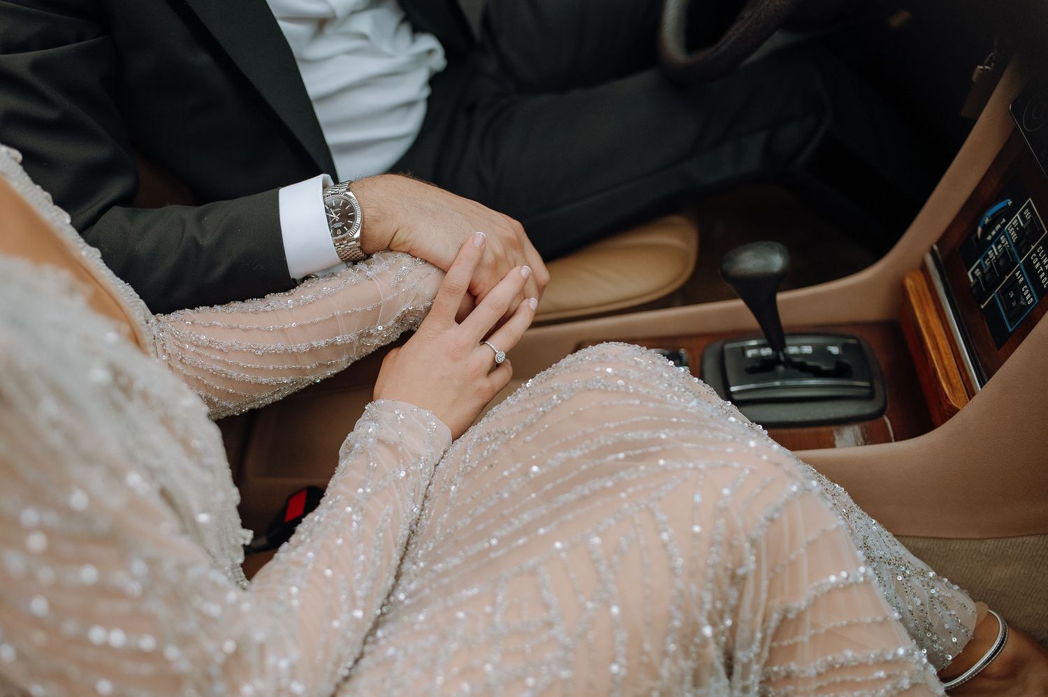 Two hands tenderly clasped together on a car seat, with one wearing formal attire and the other in a sparkly light dress.