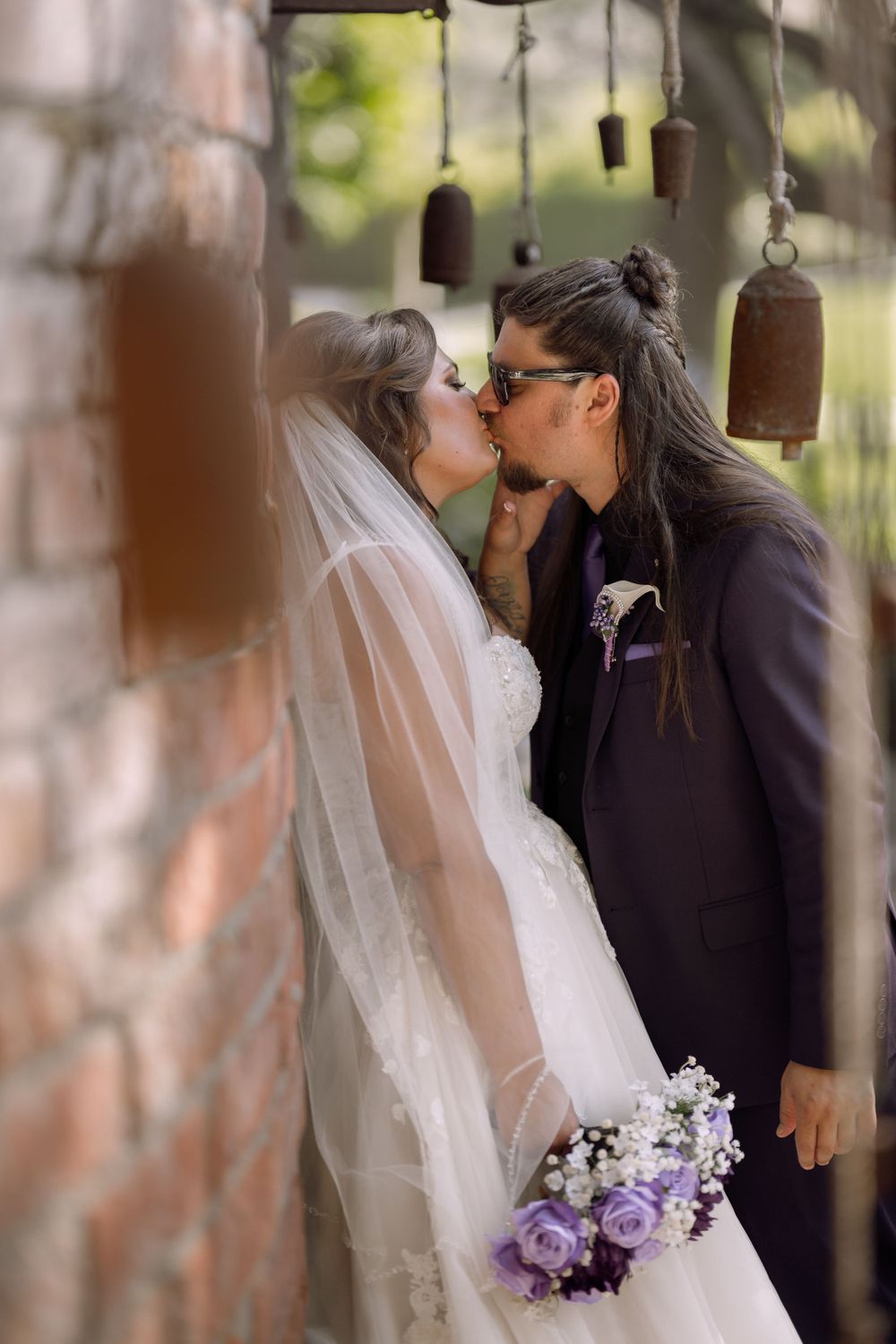 Bride and groom kissing against a brick wall with hanging ceramic bells, captured with a low depth of field and romantic vibes.