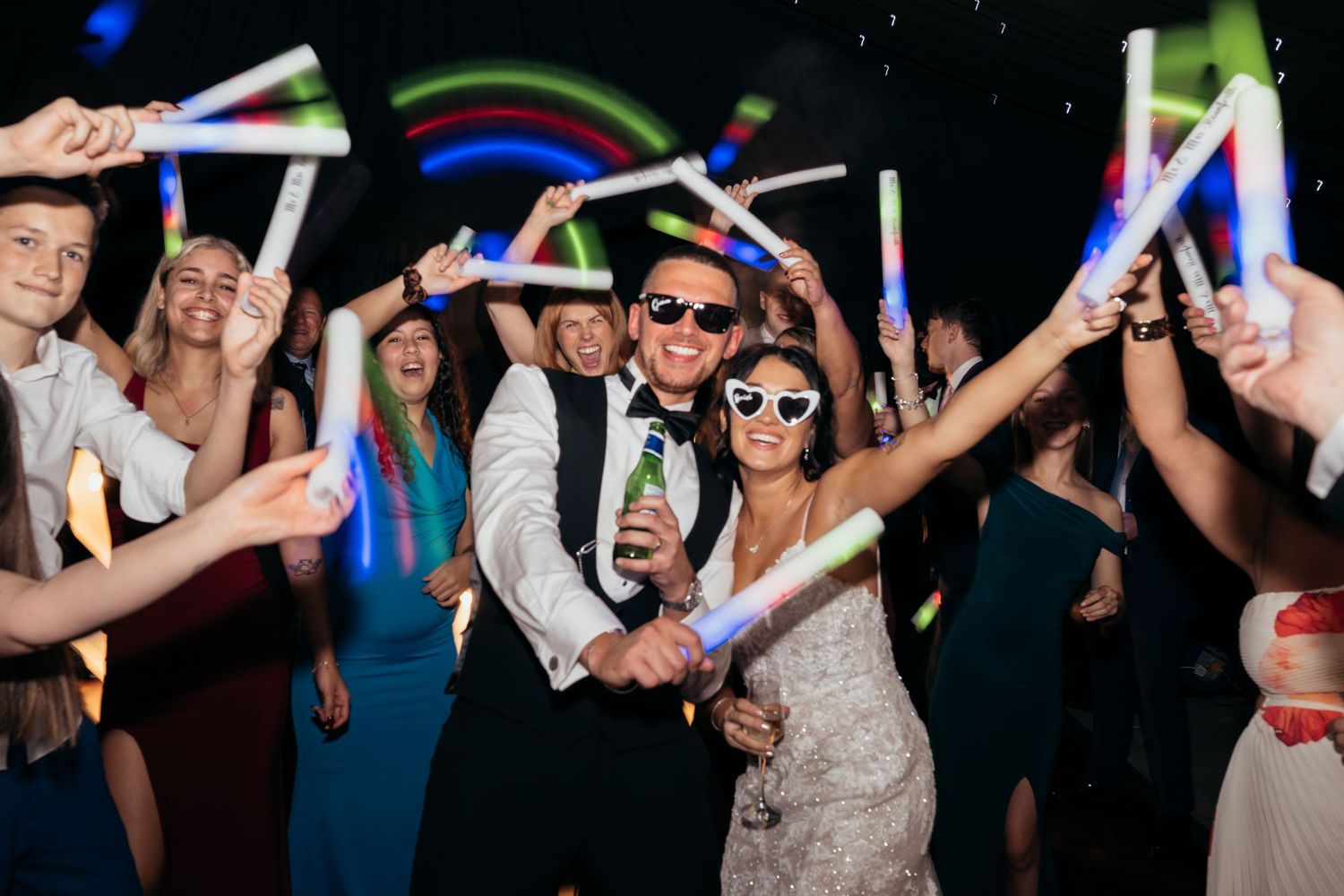 Wedding guests dance and celebrate on a dark dance floor with glowing blue lights and raised hands.