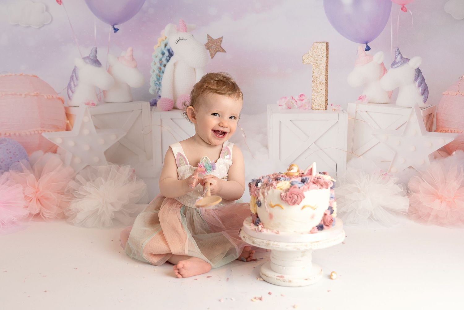 A baby's first birthday cake smash photo session with pink and purple balloons and decorations against a white backdrop.