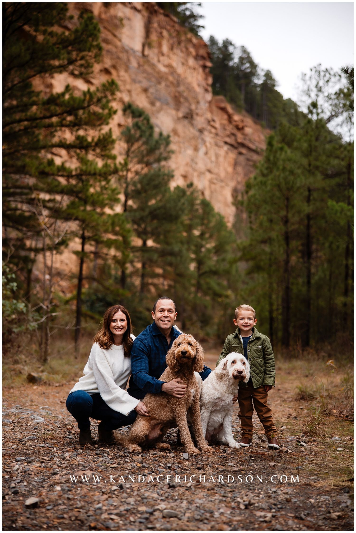 A group sits together with their golden doodle and white dogs against a rocky mountain backdrop in an outdoor setting.