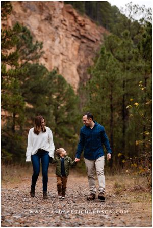 A family walks together on a dirt path with red rock cliffs and pine trees in the background.
