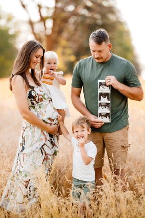 Family of four poses in golden field at sunset while looking at ultrasound photos.