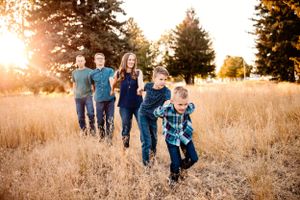 Family running together through golden field at sunset with evergreen trees in background.