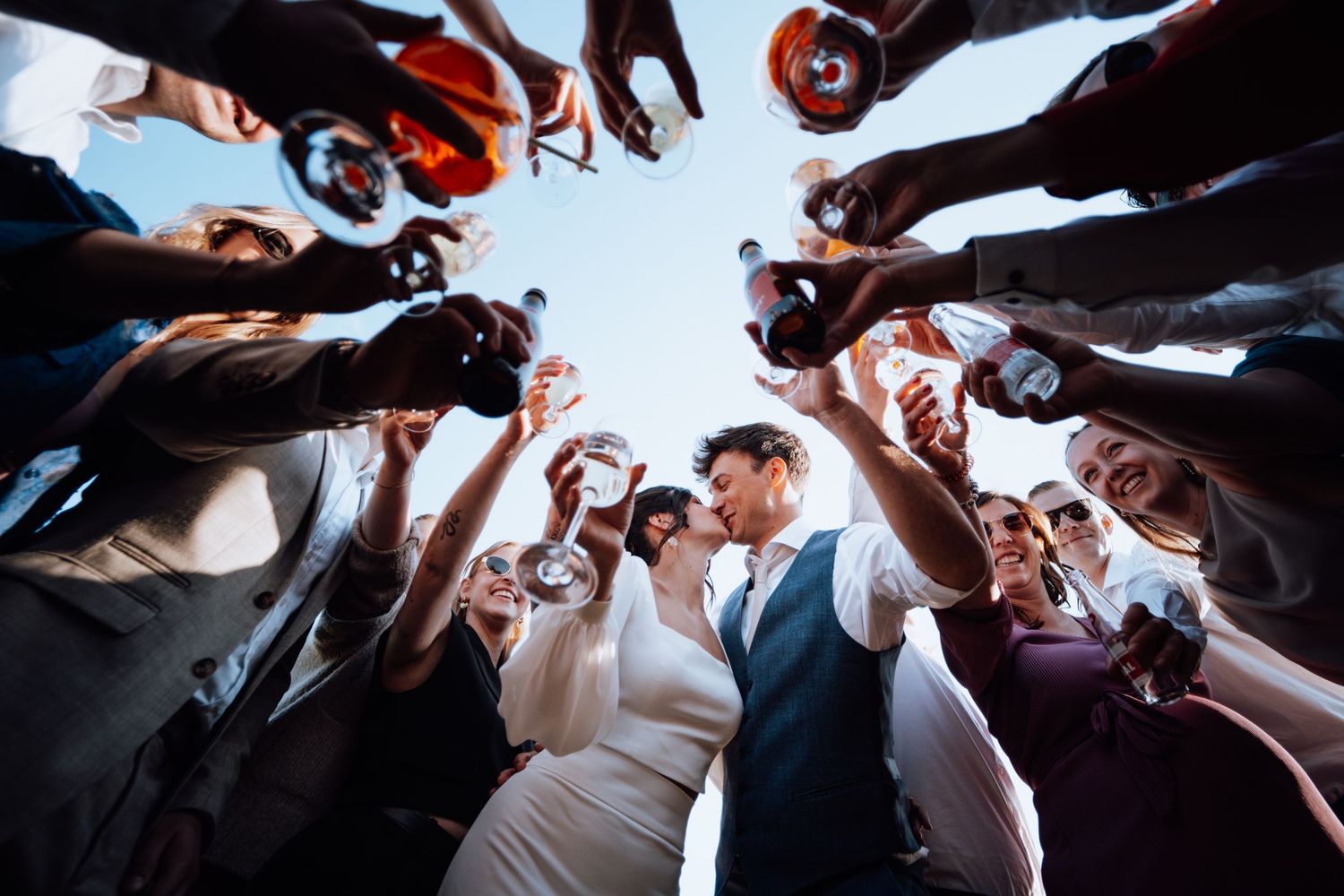 Het trouwkoppel, van onderen gezien, omringd door een feestelijke cirkel van gasten die het glas heffen op de receptie.