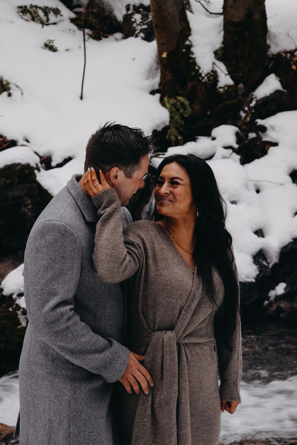 Couple in matching gray coats sharing a romantic moment in a snowy winter landscape.