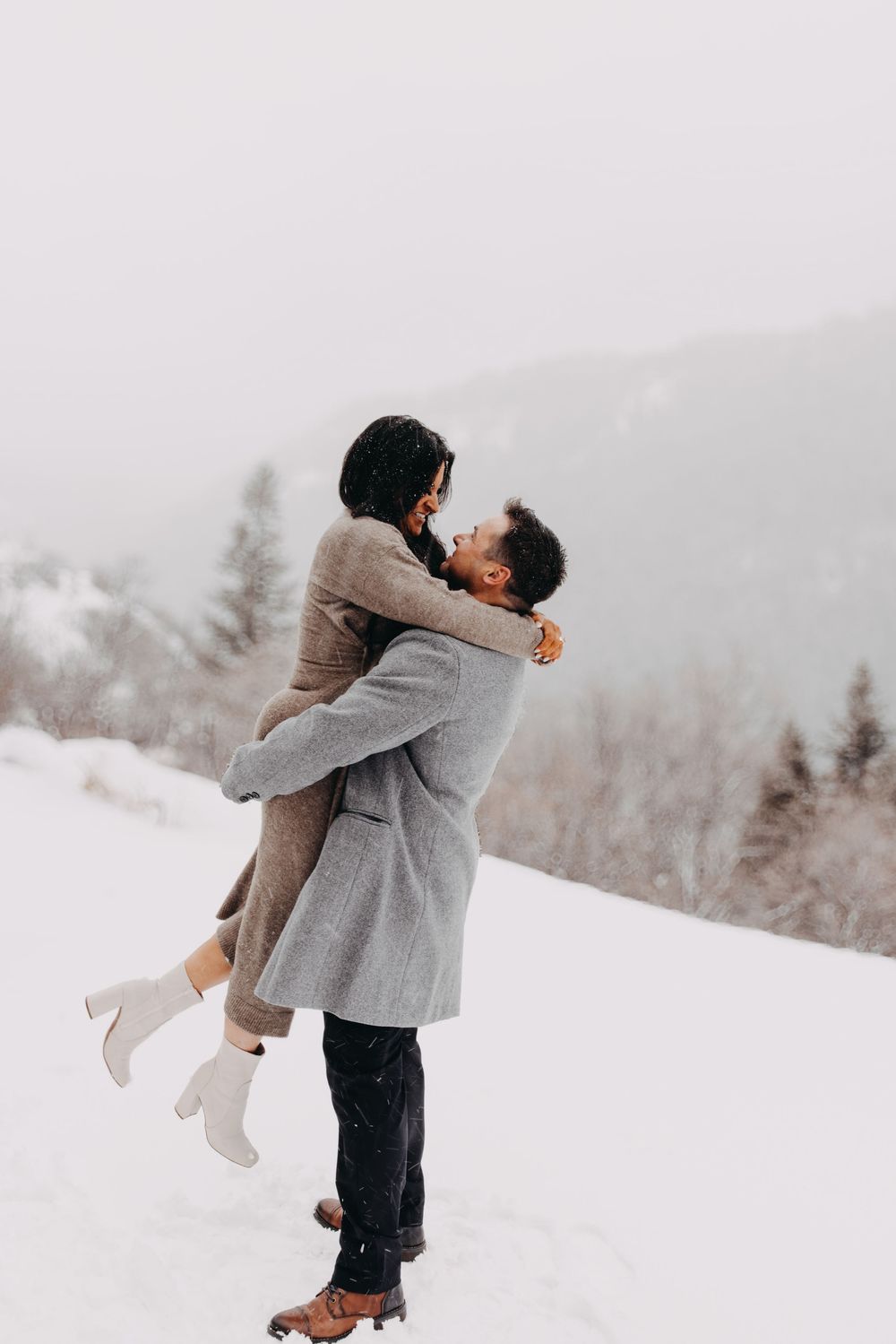 Couple embracing in snowy winter landscape, man lifting woman in gray coat, romantic outdoor photo.