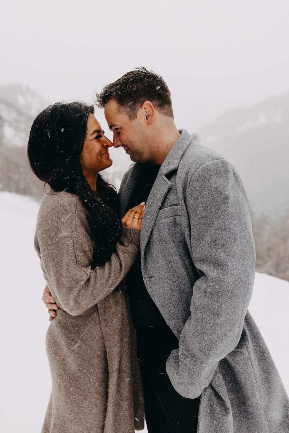Couple sharing a romantic moment in snowy winter landscape, wearing wool coats, with misty mountains in background.