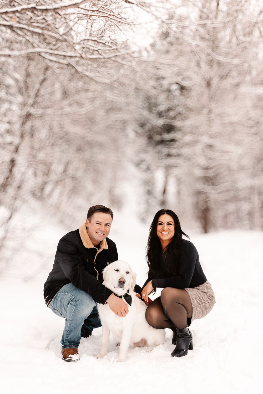 Couple crouching with white Labrador in snowy winter forest, wearing black outfits, smiling at camera.