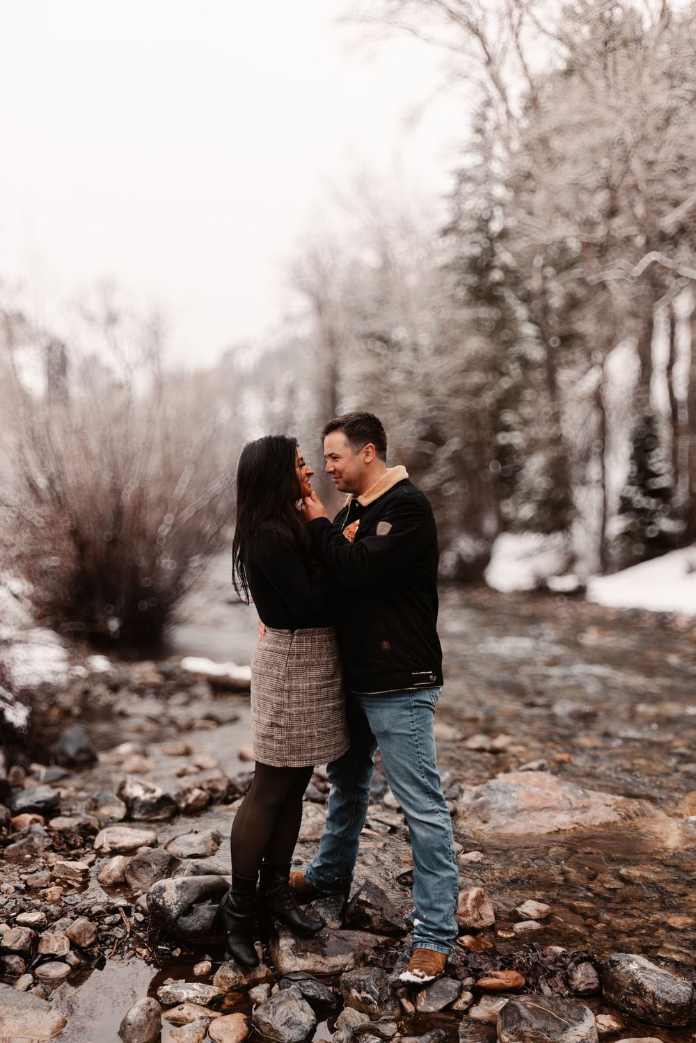 Couple embracing and laughing by a rocky creek in a snowy winter landscape, wearing cozy fall outfits.