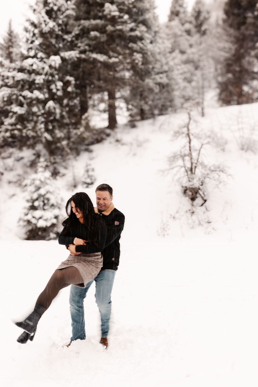 Couple playfully dancing in snowy winter landscape surrounded by snow-covered pine trees.