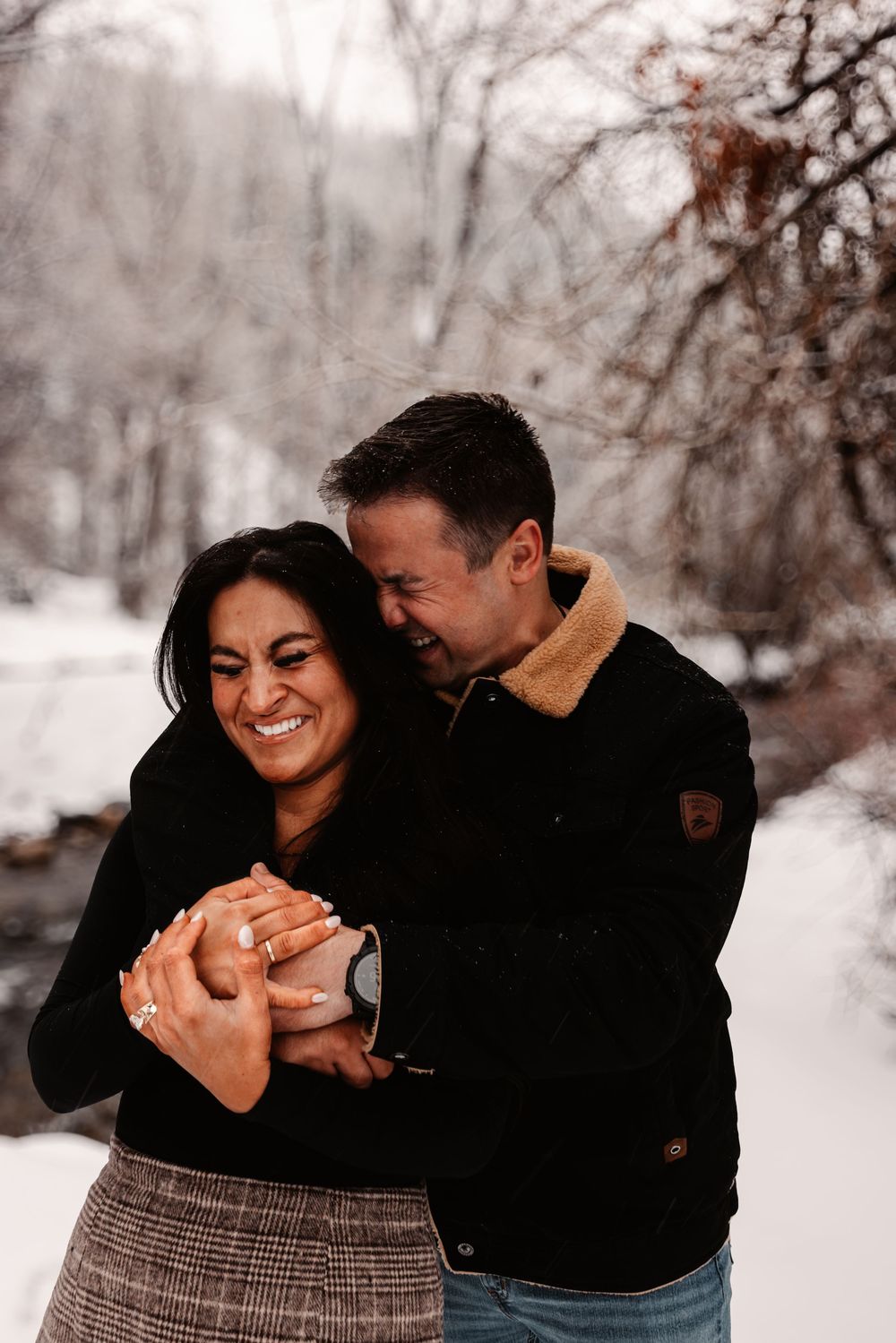 Couple laughing and embracing in a snowy winter landscape, woman wearing engagement ring, both dressed in dark winter coats.