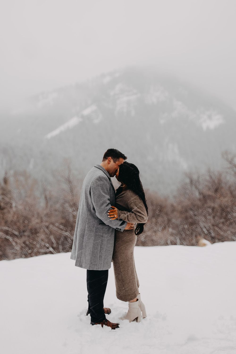 Couple kissing in snowy mountain landscape, wearing elegant winter coats, romantic outdoor engagement photo session.