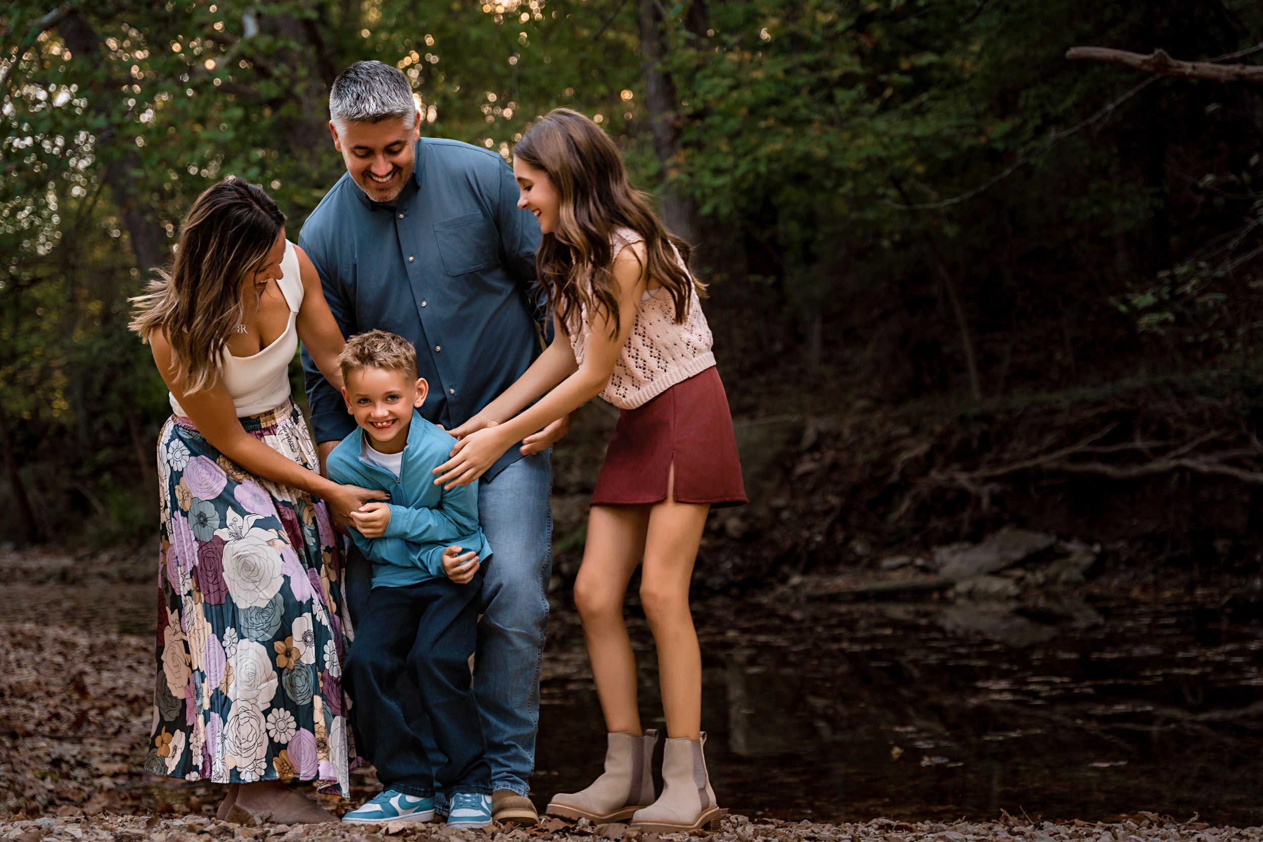 A family shares a joyful moment together while standing near a wooded creek bank during an outdoor photo session.