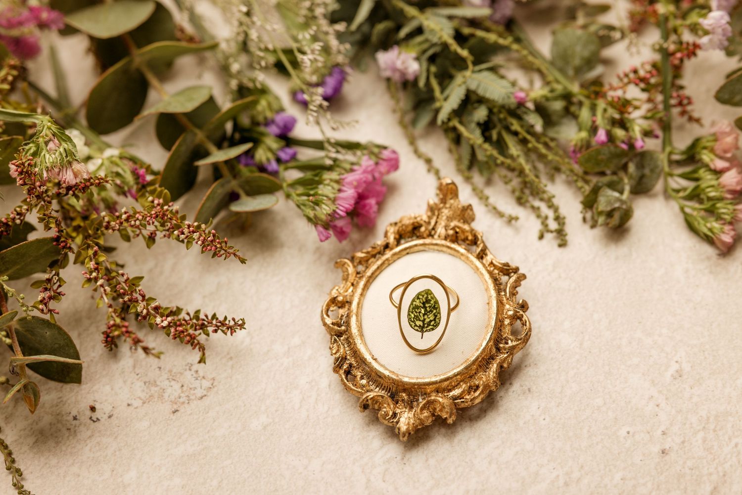 Gold ornate ring with green gemstone displayed on white surface surrounded by wildflowers and eucalyptus.