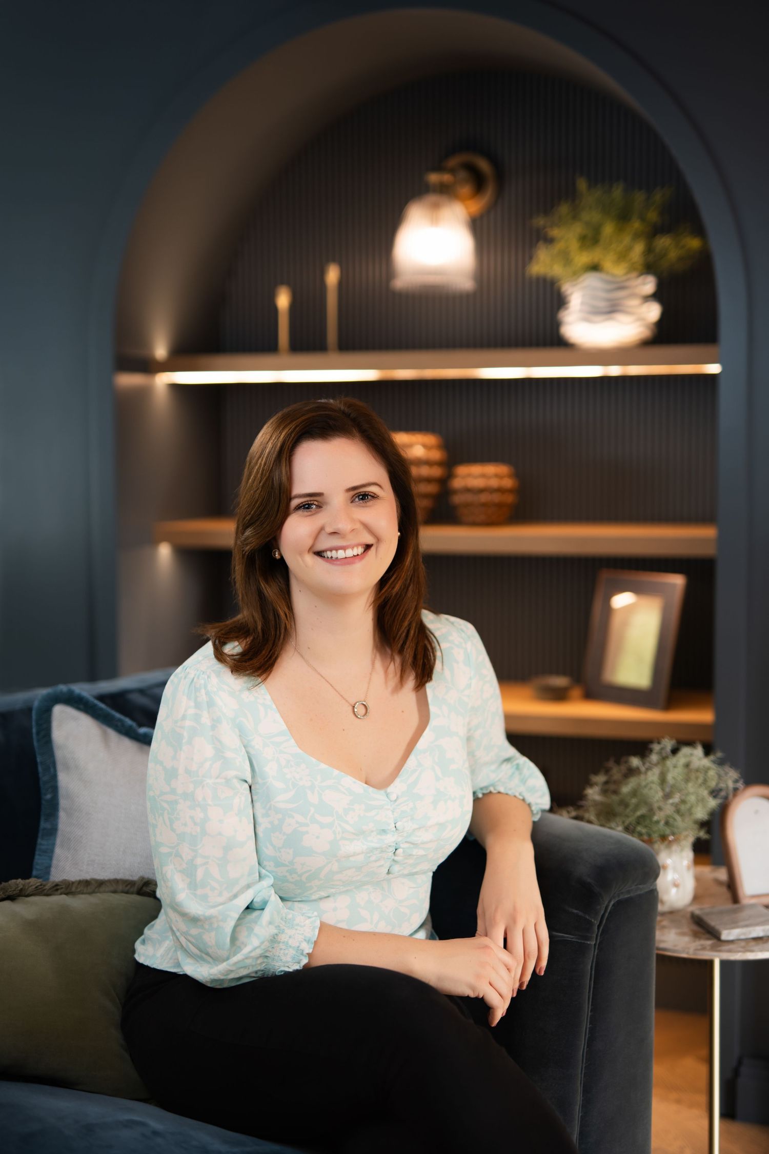 A woman in a white floral dress poses on a dark couch in a dimly lit room with modern decor.