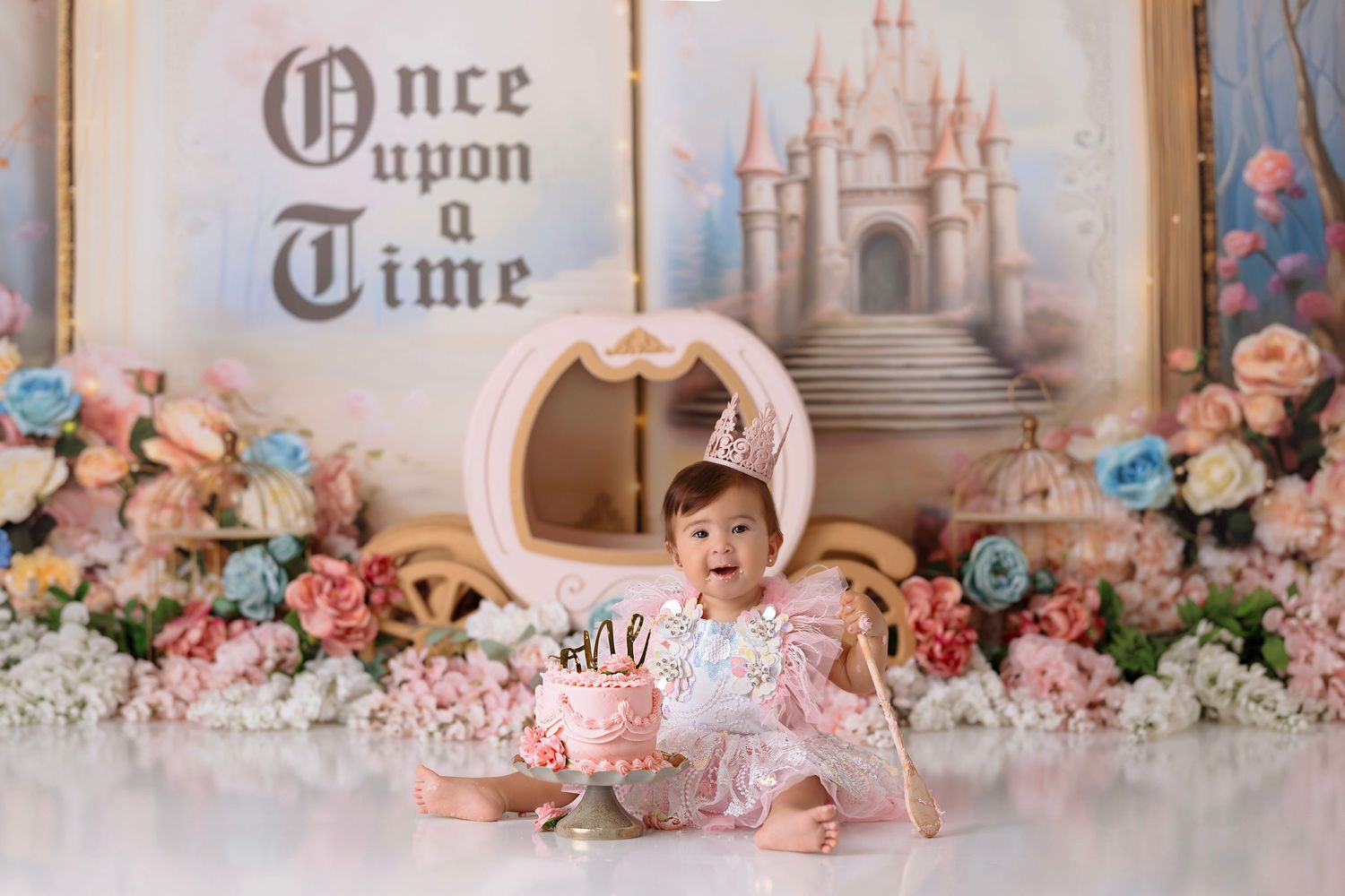 A baby sits in a dreamy princess-themed set with a white carriage, pink flowers, and a fairytale castle backdrop.