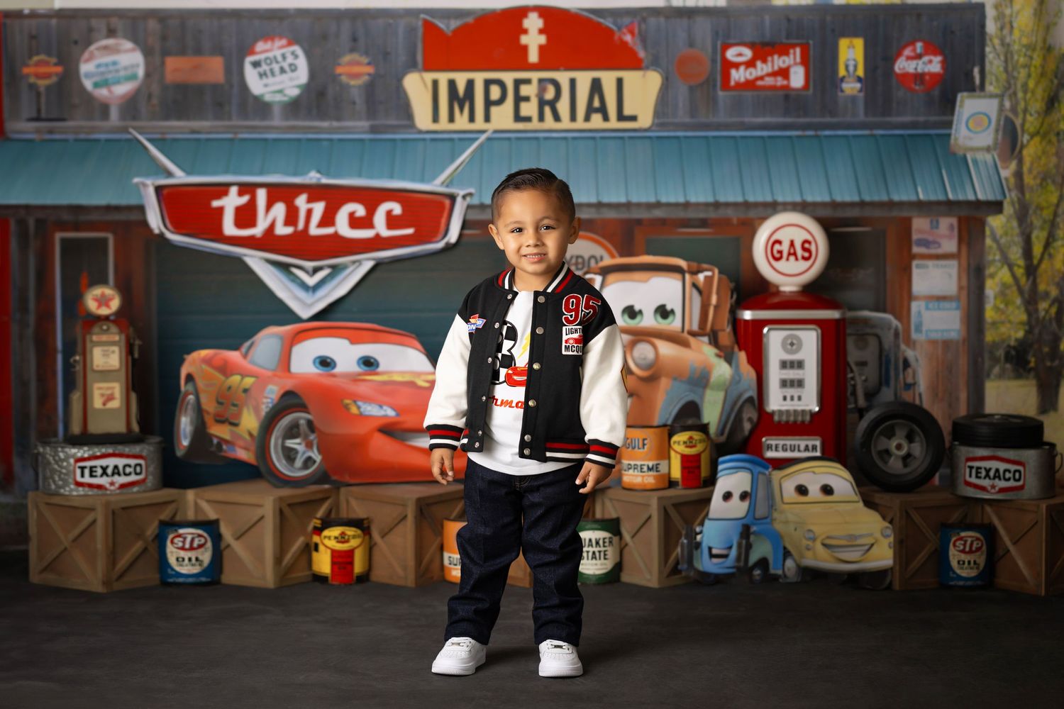 A child stands in front of a Cars-themed backdrop with vintage gas station decorations and cartoon vehicle artwork.