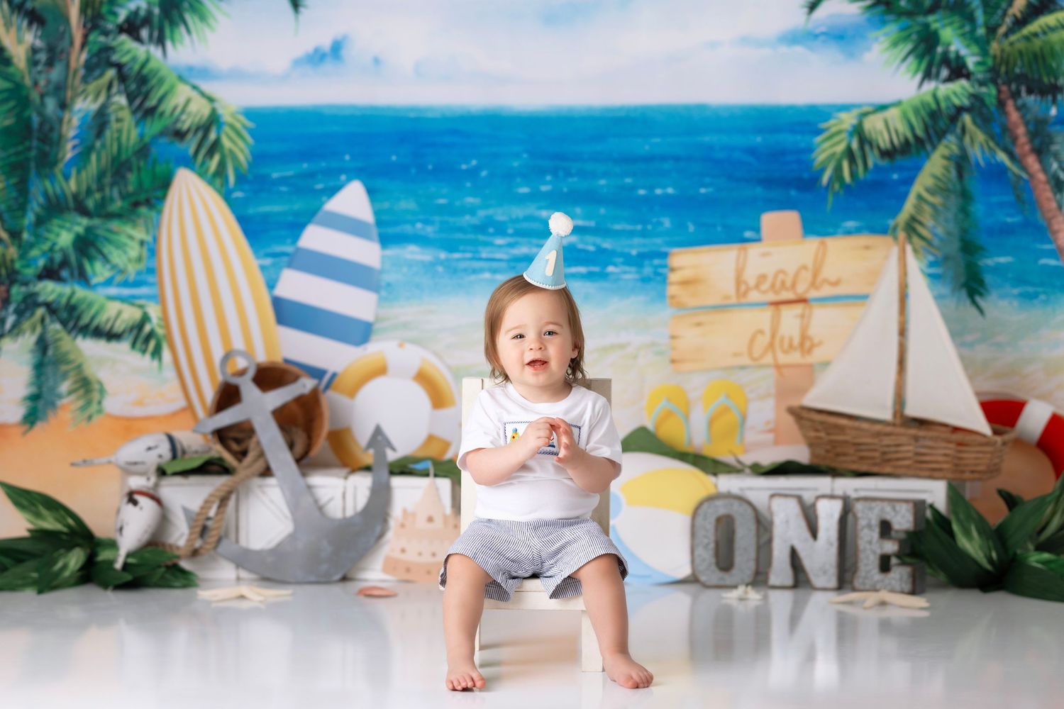 Beach themed first birthday photo setup with surfboards, palm trees, and ocean backdrop decorations.