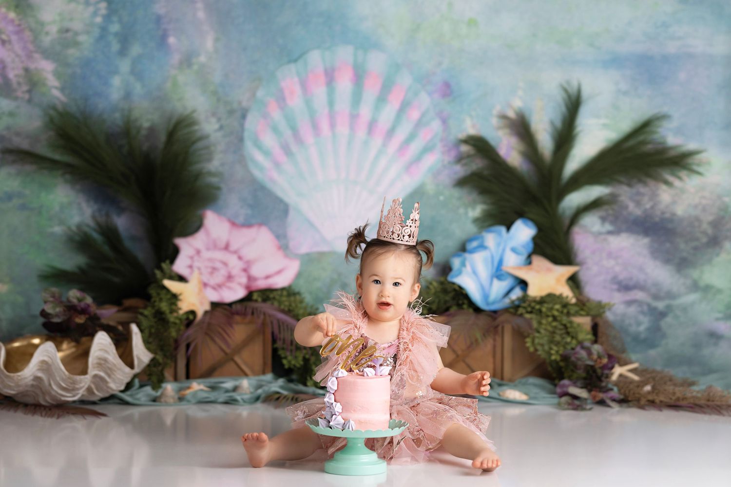 A baby in a pink tutu dress sits by a first birthday cake with mermaid-themed decorations and seashell backdrop.