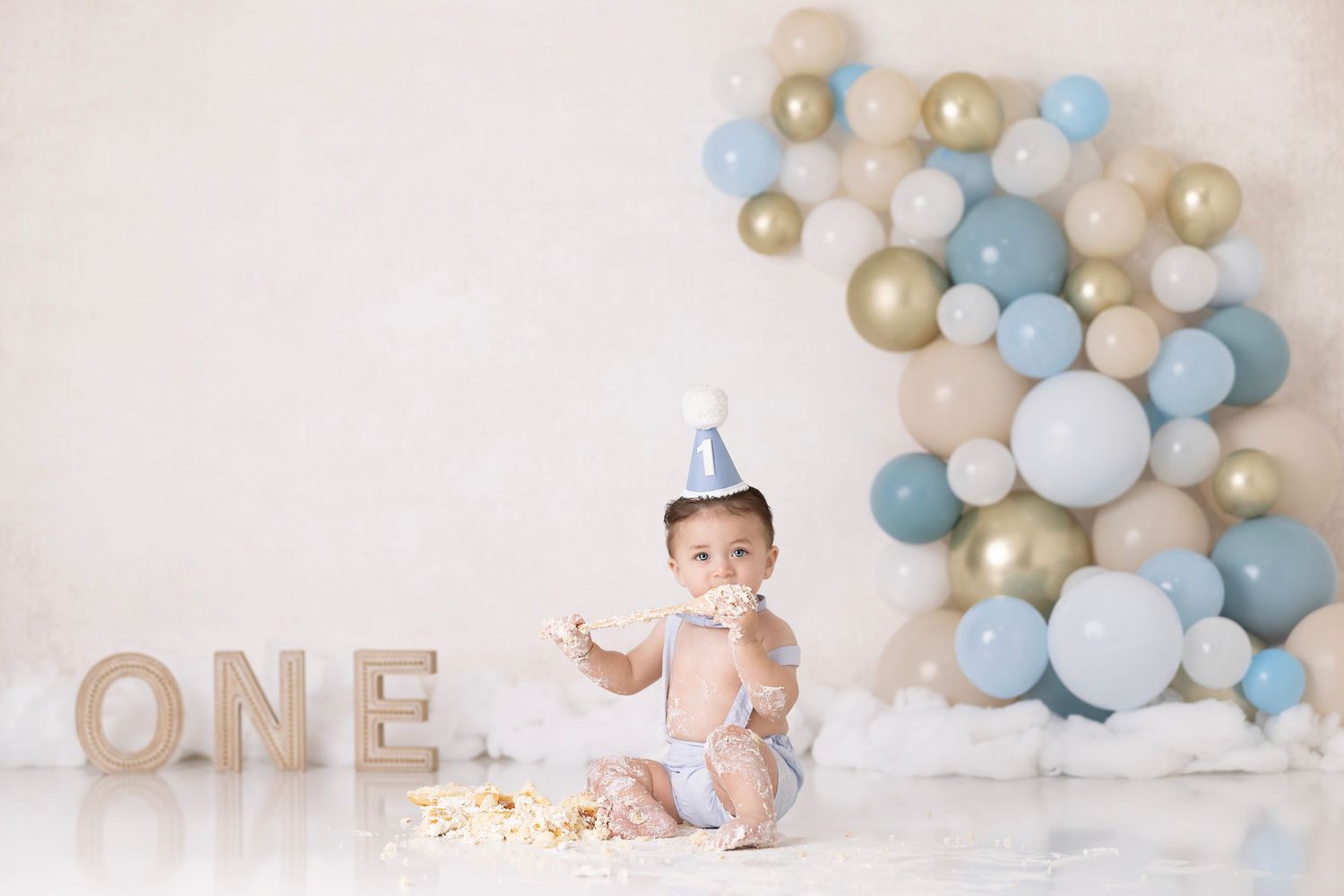 Baby in diaper and party hat sits by ONE text letters with blue and gold balloon arch during cake smash photo session.