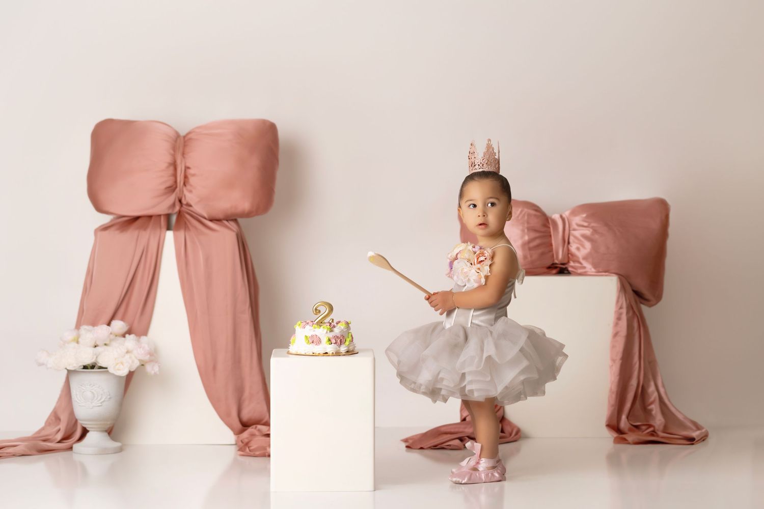 A small dancer in a tutu spins near a decorative pink bow backdrop and pedestal with a golden crown.