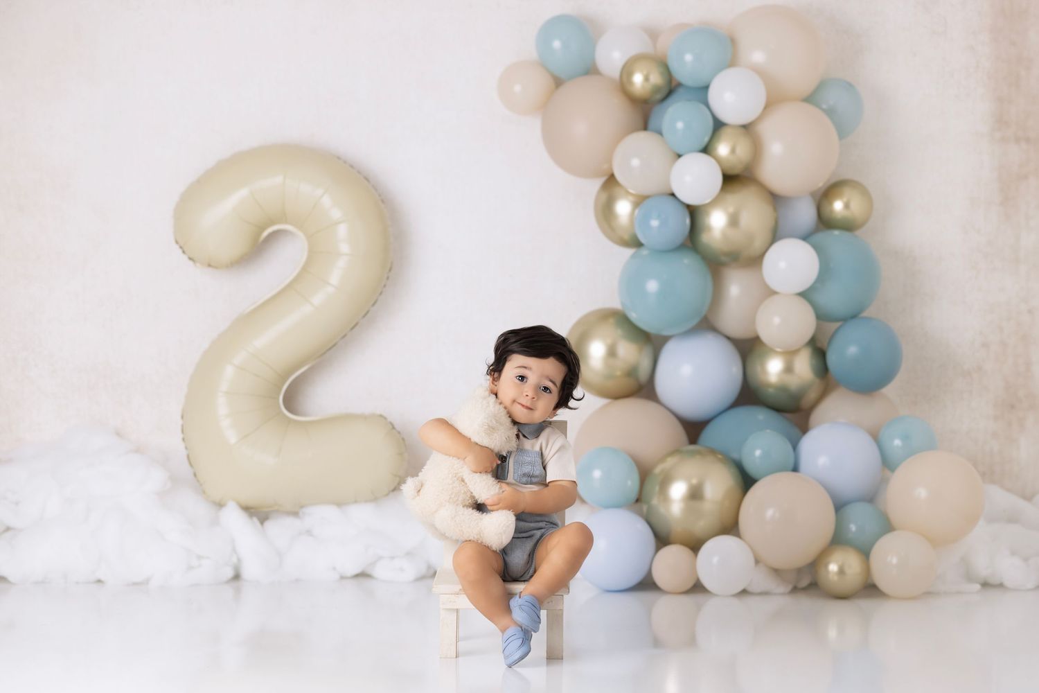 Birthday celebration setup with large number 2 balloon and blue, white and gold balloon arch against white backdrop.