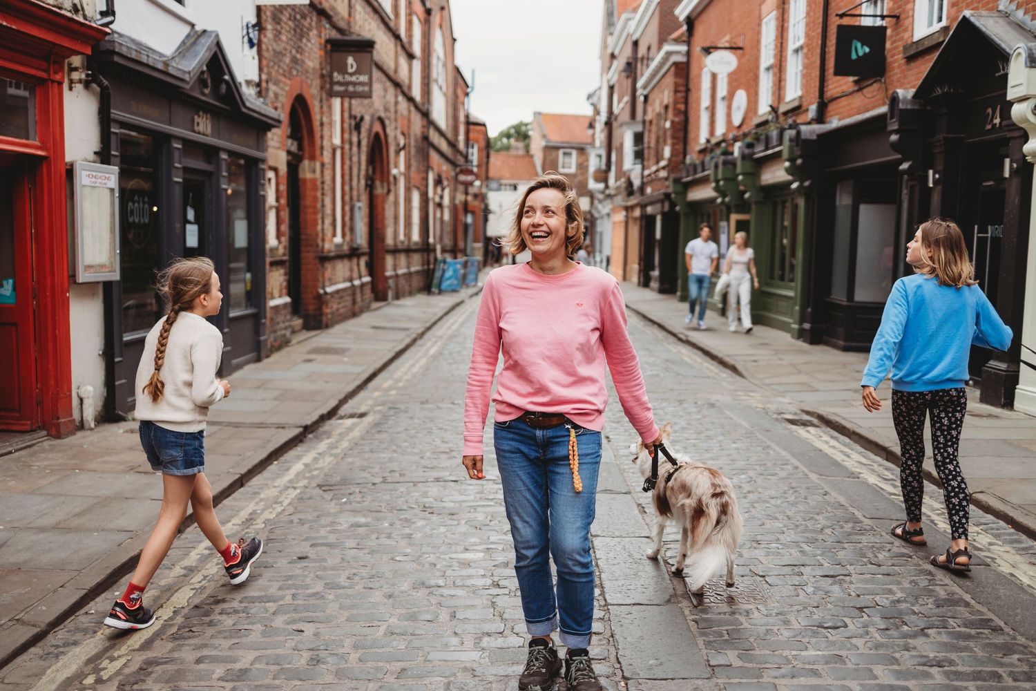 A small dog and people walking down a charming cobblestone street lined with historic brick buildings.