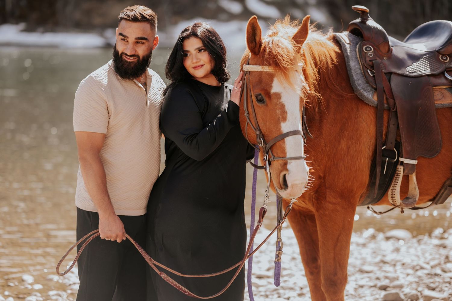 A couple poses with a saddled brown horse against a wintry landscape.