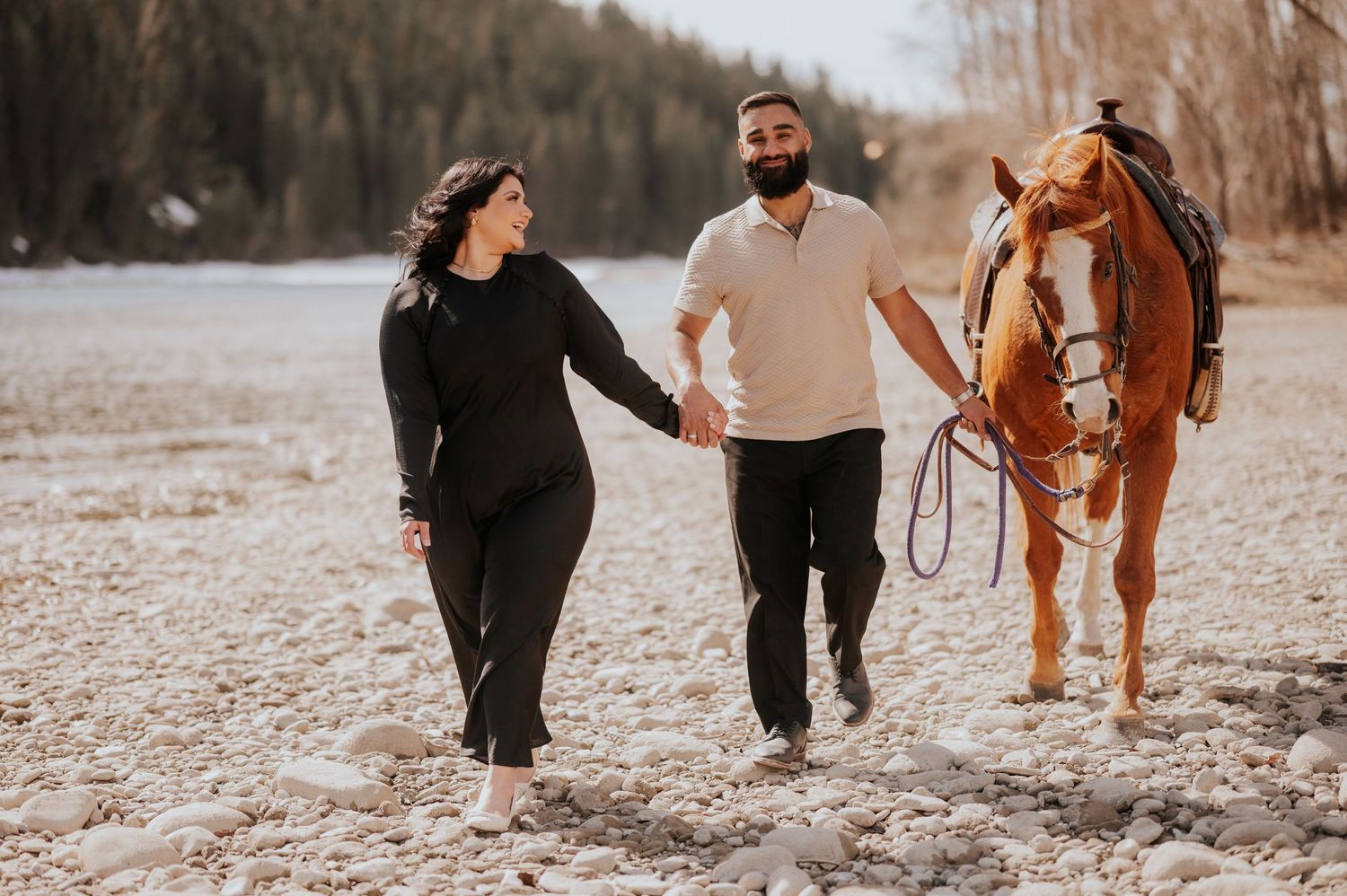 A couple walking along a beach with their brown horse following behind.