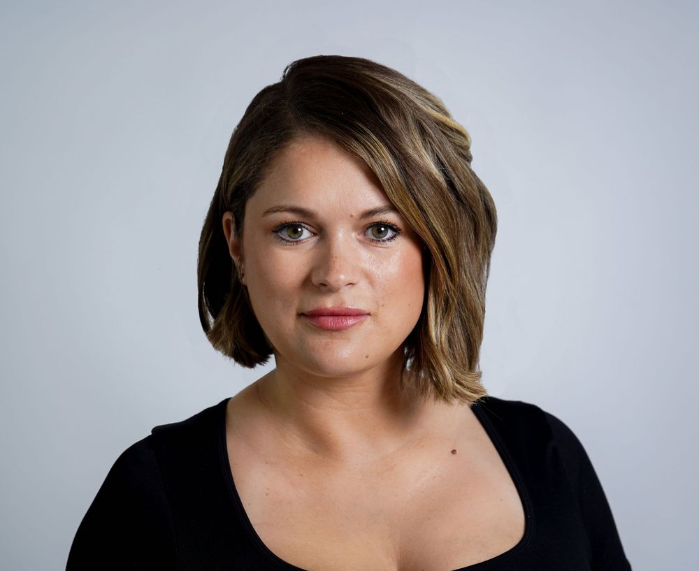 Professional headshot with short brown hair wearing a black top against a light gray background.