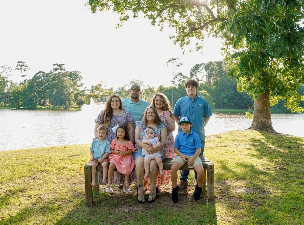 A group sits together outdoors on a wooden bench against a lush green backdrop near a serene waterfront setting.