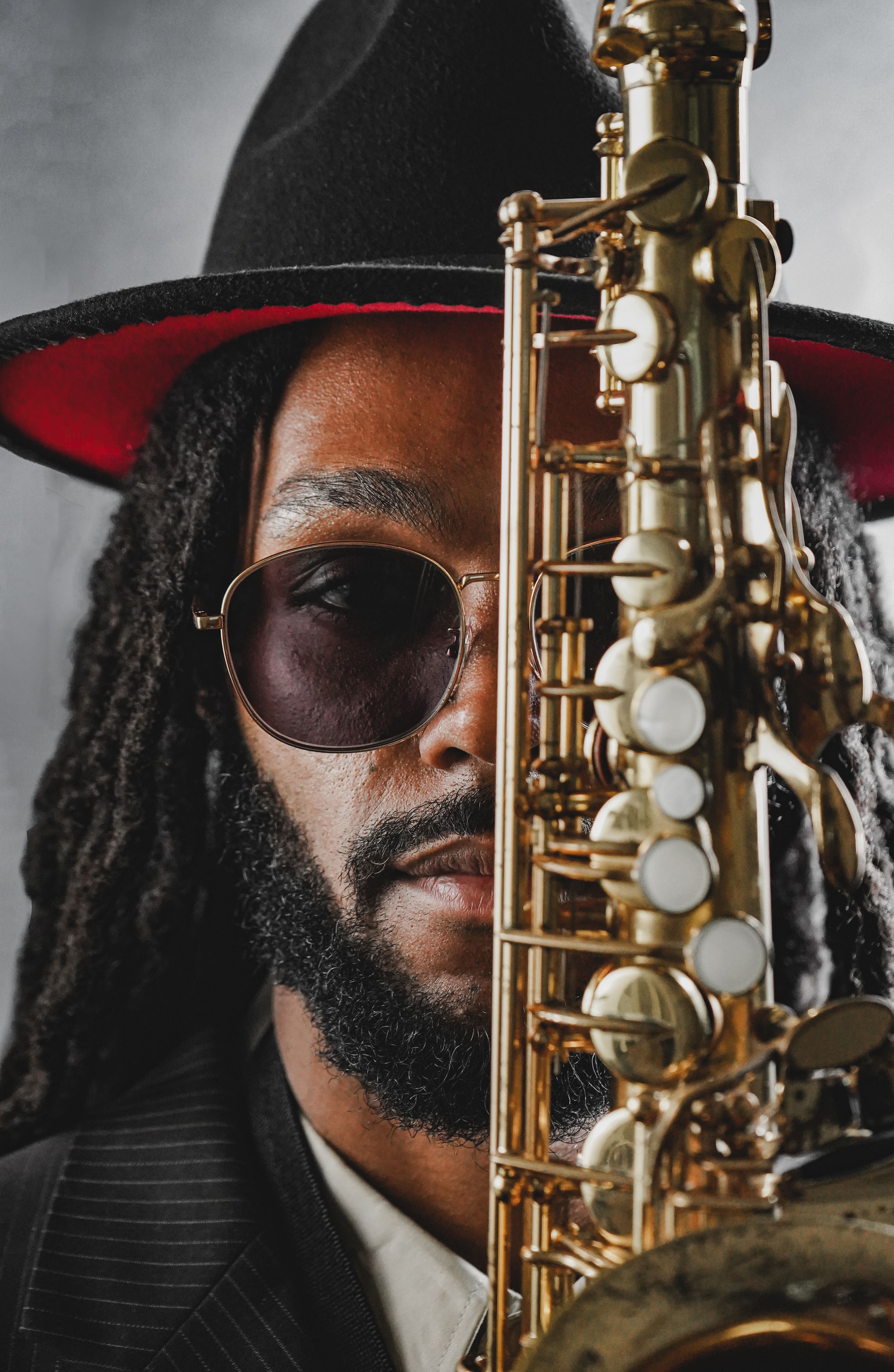 A jazz musician with dreadlocks and sunglasses holds a gold soprano saxophone beside his face, wearing a wide-brim hat.