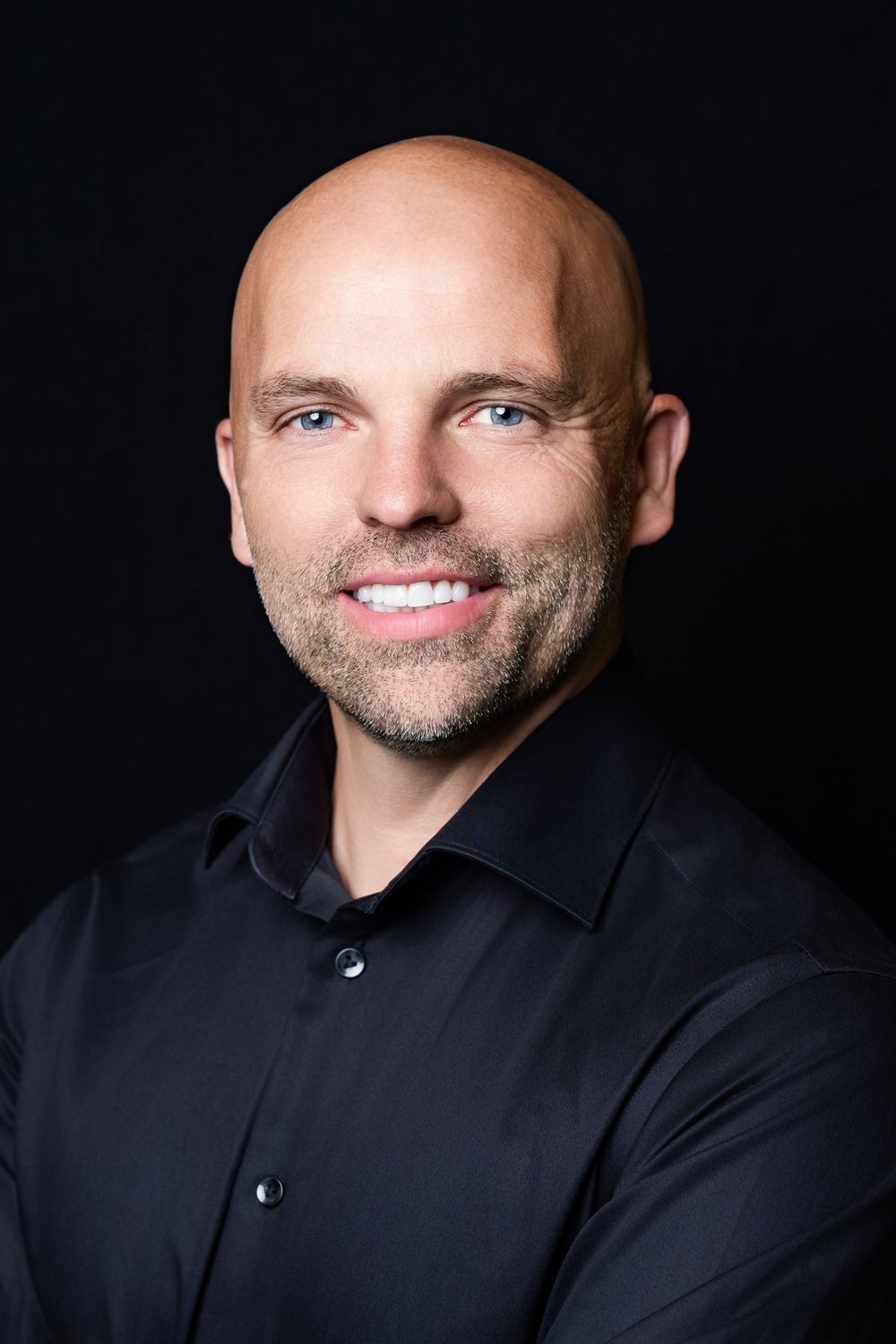 Professional headshot portrait of a smiling person with no hair wearing a black button-down shirt against a dark background.