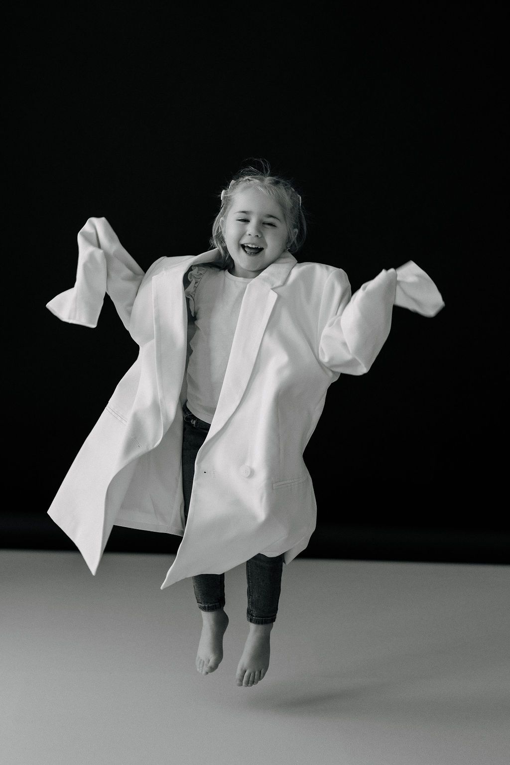 Joyful child jumping in oversized white dress shirt against black background.